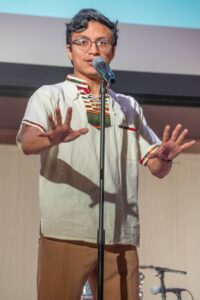 Matthew Marroquín speaking passionately into a microphone on stage, wearing glasses and a light-colored shirt.